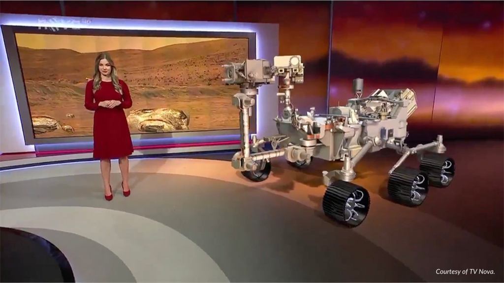 A TV Nova newscaster stands next to an AR version of the Perseverance rover.